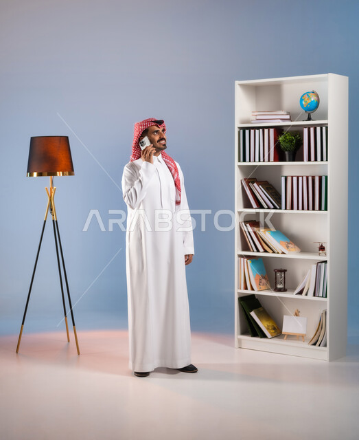 إجراء مكالمة هاتفية عبر الجوال, بورتريه لرجل عربي خليجي سعودي يرتدي الثوب التقليدي والشماغ يستخدم الهاتف المحمول, التواصل مع الأقارب والأصدقاء عبر وسائل التواصل الاجتماعي, الاستمتاع بتبادل الأحاديث والأخبار, صورة الجسم بالطول الكامل, خلفية ملونة