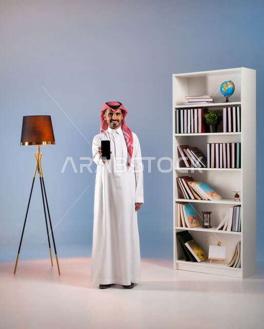 النظر إلى الكاميرا بإيماءات السرور, عرض تطبيقات وخدمات الهاتف المحمول, بورتريه لشاب عربي خليجي سعودي يرتدي الثوب التقليدي والشماغ يحمل الجوال بشاشة سوداء فارغة, استخدام الأجهزة والتقنيات الحديثة, صورة الجسم بالطول الكامل, خلفية ملونة