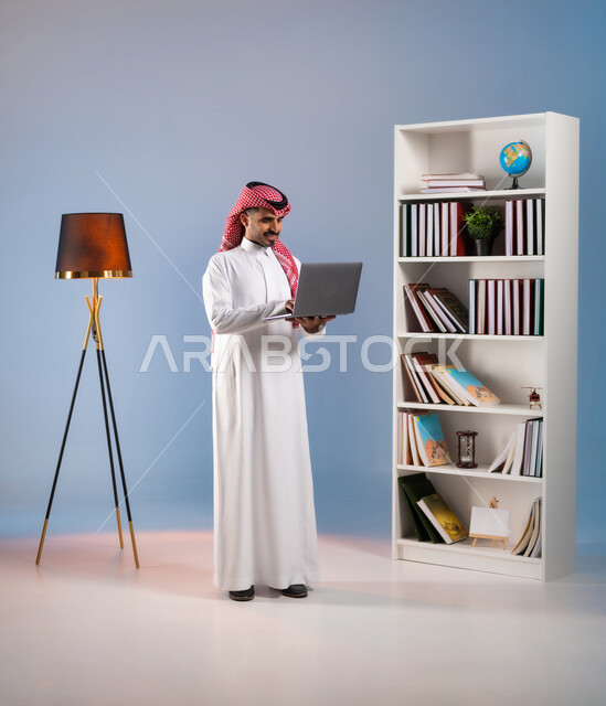 إدراة وانجاز المهام عبر الانترنت, الدردشة والمحادثة مع الأصدقاء عبر السوشال ميديا, تصفح مواقع التواصل الاجتماعي, بورتريه لرجل عربي خليجي سعودي مبتسم يرتدي الثوب التقليدي والشماغ يعمل على الحاسب المحمول, صورة الجسم بالطول الكامل, خلفية ملونة