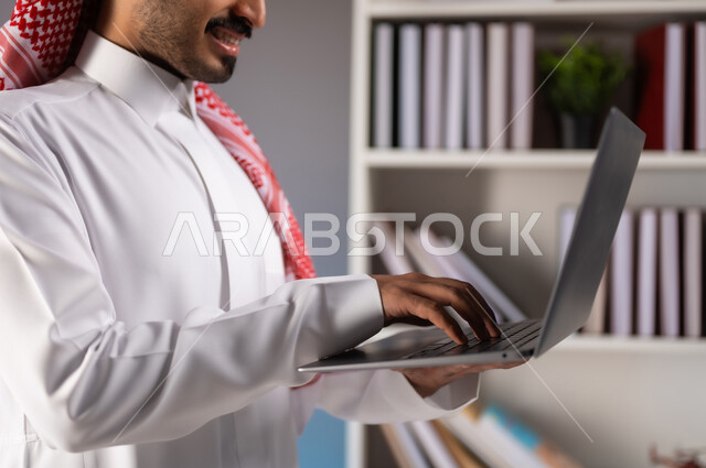 تصفح مواقع التواصل الاجتماعي, بورتريه مقرب لرجل عربي خليجي سعودي يرتدي الثوب التقليدي والشماغ يعمل على الحاسب المحمول, إدراة وانجاز المهام عبر الانترنت, الدردشة والمحادثة مع الأصدقاء عبر السوشال ميديا, خلفية ملونة