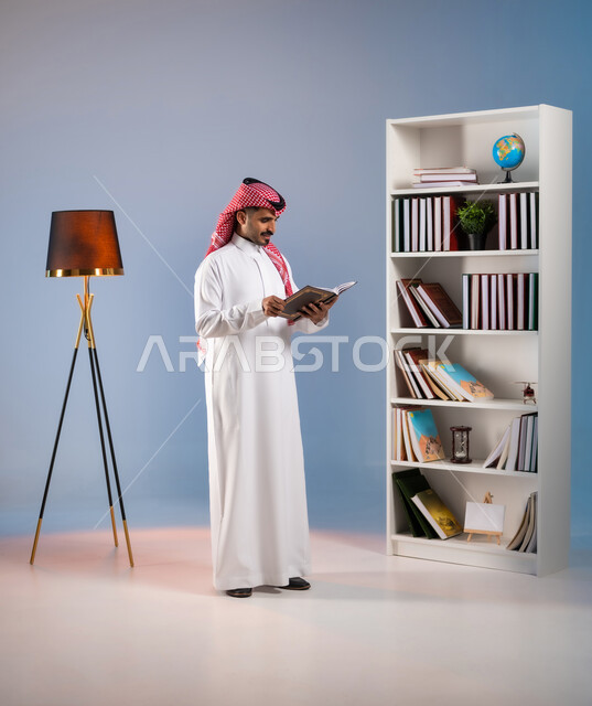 قراءة القصص والروايات الممتعة, بورتريه لرجل عربي خليجي سعودي يرتدي الثوب التقليدي والشماغ يمسك بيده كتاب, تنمية الذخيرة الثقافية, قضاء أوقات الفراغ بممارسة هواية المطالعة, مفهوم الثقافة والتعلم الذاتي, صورة الجسم بالطول الكامل, خلفية ملونة