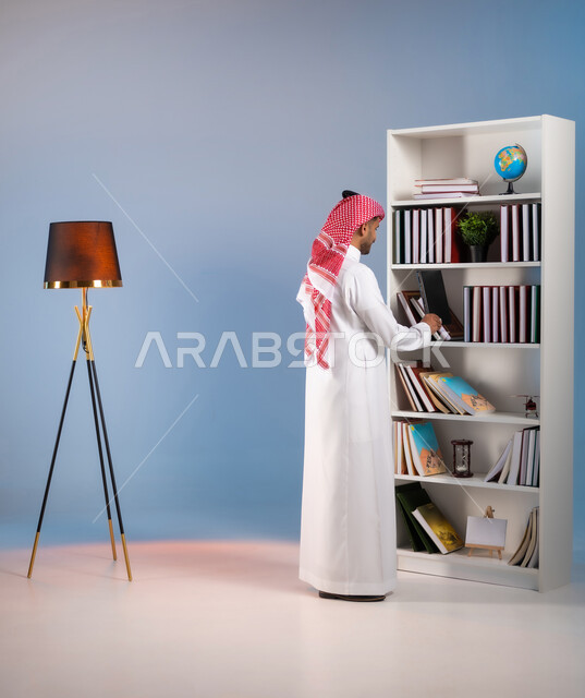 الاستمتاع بممارسة الهوايات المفيدة, مفهوم القراءة والثقافة, تنمية الذخيرة العلمية, محبي وهواة المطالعة في المملكة, بورتريه من الخلف لرجل عربي خليجي سعودي يرتدي الثوب التقليدي والشماغ يقوم باقتناء الكتاب من المكتبة, صورة الجسم بالكامل, خلفية ملونة