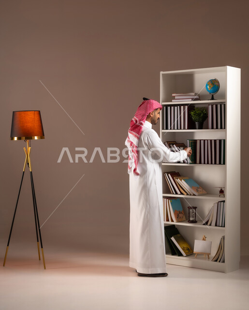 مفهوم القراءة والثقافة, تنمية الذخيرة العلمية, محبي وهواة المطالعة في المملكة, الاستمتاع بممارسة الهوايات المفيدة, بورتريه من الجانب لرجل عربي خليجي سعودي يرتدي الثوب التقليدي والشماغ يقوم باقتناء الكتاب من المكتبة, صورة الجسم بالكامل, خلفية بيج