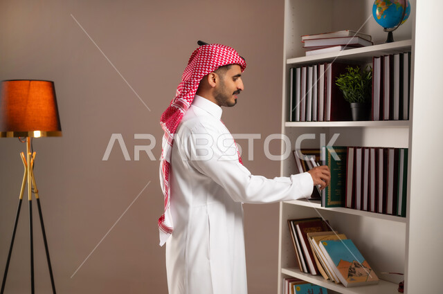 تنمية الذخيرة العلمية, اقتناء واختيار الكتاب من المكتبة, مفهوم القراءة والثقافة, محبي وهواة المطالعة في المملكة, الاستمتاع بممارسة الهوايات المفيدة, بورتريه من الجانب لرجل عربي خليجي سعودي يرتدي الثوب التقليدي والشماغ يقوم بأخذ الكتاب, خلفية بيج