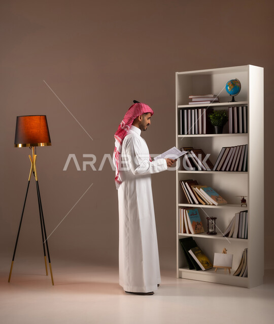 ممارسة الهوايات الممتعه والمفيدة في وقت الفراغ, قراءة الكتب التعليمية التثقيفة, بورتريه من الجانب لرجل عربي خليجي سعودي يرتدي الزي التقليدي يمسك بيده الكتاب, مفهوم التعلم الذاتي, اقتناء واختيار الكتب من المكتبة المنزلية. صورة الجسم بالطول الكامل, خلفية بيج