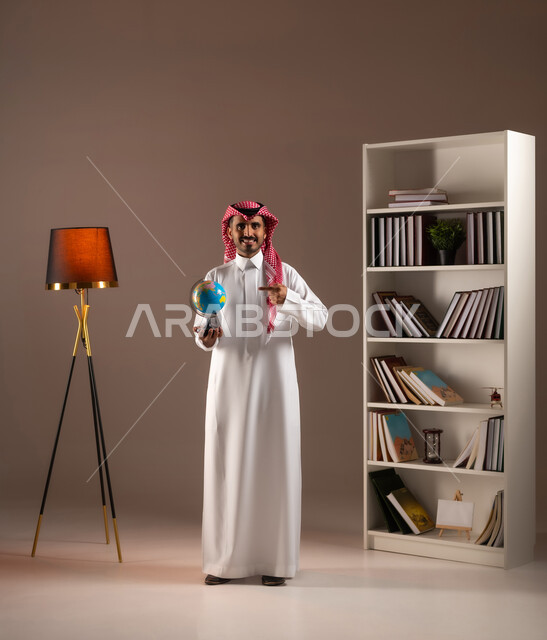 مفهوم الرحلات والجولات السياحية, البحث والاستكشاف حول العالم, النظر إلى الكاميرا والاشارة إلى مكان معين على الخريطة, بورتريه لرجل عربي خليجي سعودي مبتسم يرتدي الثوب التقليدي والشماغ يحمل بيده مجسم الكرة الارضية, صورة الجسم بالكامل, خلفية بيج