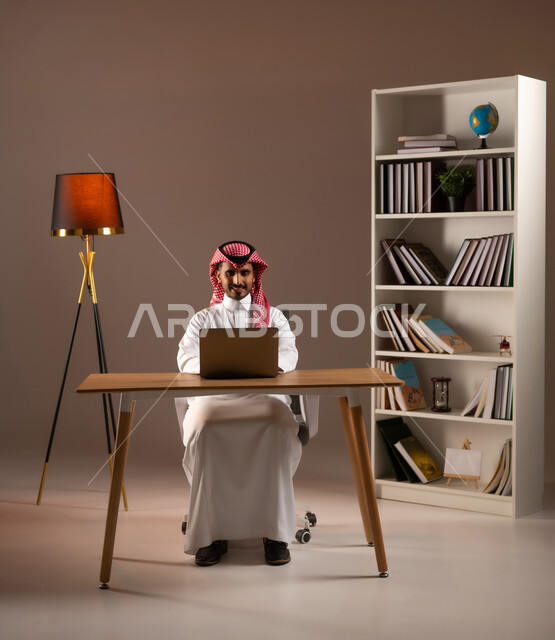 التسوق الالكتروني والشراء عبر التطبيقات والتقنيات الحديثة, بورتريه لرجل عربي خليجي سعودي يرتدي الثوب التقليدي والشماغ يعمل على الحاسب المحمول, استخدام الأجهزة المتطورة في مكاتب المملكة, إدراة الأعمال عن بعد, انجاز المهام عبر الانترنت, خلفية بيج