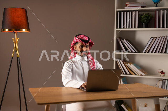 إدراة الأعمال عن بعد, انجاز المهام عبر الانترنت, التسوق الالكتروني والشراء عبر التطبيقات والتقنيات الحديثة, بورتريه لرجل عربي خليجي سعودي يرتدي الثوب التقليدي والشماغ يعمل على الحاسب المحمول, استخدام الأجهزة المتطورة في مكاتب المملكة, خلفية بيج