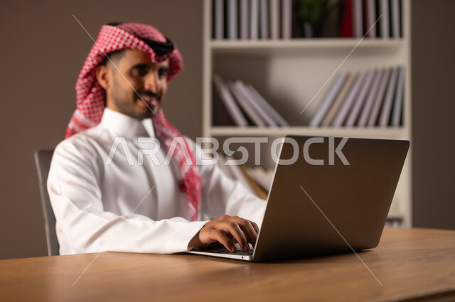 انجاز المهام عبر الانترنت, التسوق الالكتروني والشراء عبر التطبيقات والتقنيات الحديثة, بورتريه مقرب لرجل عربي خليجي سعودي يرتدي الثوب التقليدي والشماغ يعمل على الحاسب المحمول, استخدام الأجهزة المتطورة في مكاتب المملكة, إدراة الأعمال عن بعد, خلفية بيج
