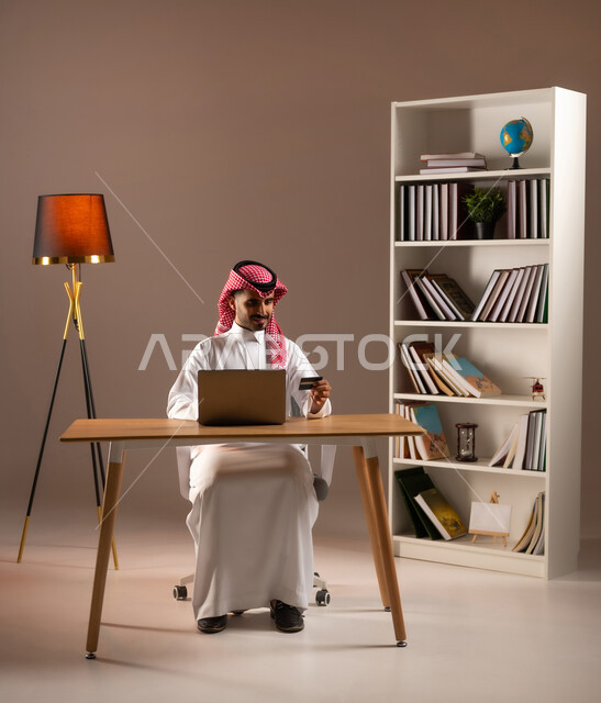 الجلوس بتركيز والعمل على الحاسب المحمول, إمكانية تسديد الفواتير بالبطاقات الإلكترونية, استخدام الأجهزة والتقنيات الحديثة, سهولة الشراء والدفع عن طريق بطاقة الإئتمان البنكية، بورتريه لرجل عربي خليجي سعودي يرتدي الزي التقليدي يمسك بيده بطاقة الصراف, خلفية بيج