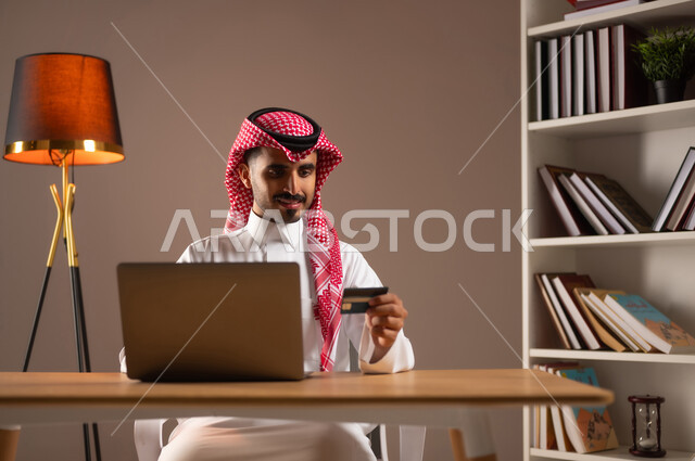 إمكانية تسديد الفواتير بالبطاقات الإلكترونية, استخدام الأجهزة والتقنيات الحديثة, سهولة الشراء والدفع عن طريق بطاقة الإئتمان البنكية، بورتريه لرجل عربي خليجي سعودي مبتسم يرتدي الثوب التقليدي والشماغ يمسك بيده بطاقة الصراف ويعمل على الحاسب المحمول, خلفية بيج