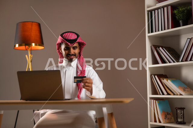 استخدام الأجهزة والتقنيات الحديثة, سهولة الشراء والدفع عن طريق بطاقة الإئتمان البنكية، بورتريه لرجل عربي خليجي سعودي مبتسم يرتدي الثوب التقليدي والشماغ يمسك بيده بطاقة الصراف ويعمل على الحاسب المحمول, إمكانية تسديد الفواتير بالبطاقات الإلكترونية, خلفية بيج