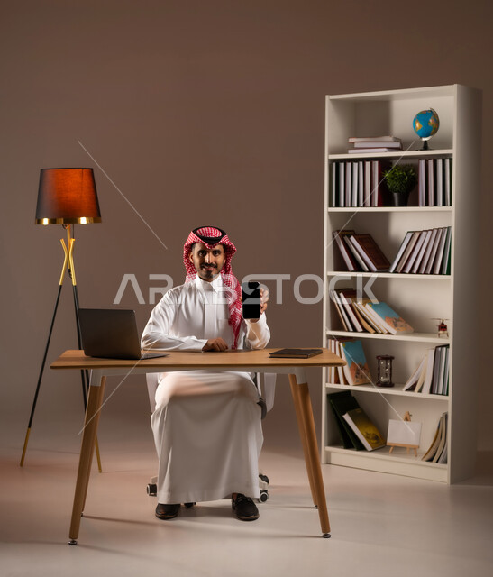 التواصل مع الأهل والأصدقاء عبر الأونلاين, عرض تطبيقات وخدمات الهاتف المحمول, بورتريه لشاب عربي خليجي سعودي يرتدي الثوب التقليدي والشماغ يحمل الجوال بشاشة سوداء فارغة, استخدام الأجهزة والتقنيات الحديثة, خلفية بيج