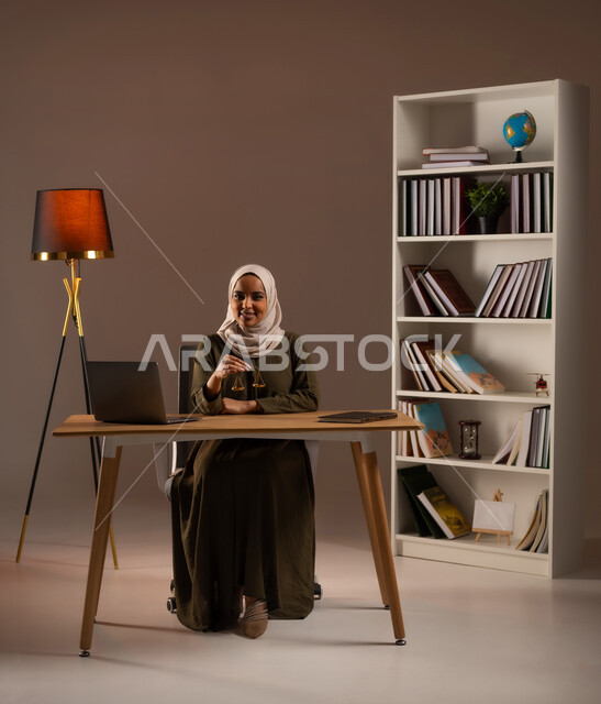 مفهوم العدالة ودراسة القضية, استخدام الأجهزة والتقنيات الحديثة في المجال القانوني, العمل في مجال القضاء والمحاماة, مهن ووظائف قانونية, دراسة القانون والحقوق, بورتريه لإمرأة عربية خليجية سعودية مبتسمة ترتدي العباءة الملونة تمسك بيدها ميزان العدل, خلفية بيج