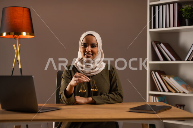 استخدام الأجهزة والتقنيات الحديثة في المجال القانوني, العمل في مجال القضاء والمحاماة, مفهوم العدالة ودراسة القضية, مهن ووظائف قانونية, دراسة القانون والحقوق, بورتريه لإمرأة عربية خليجية سعودية مبتسمة ترتدي العباءة الملونة تمسك بيدها ميزان العدل, خلفية بيج