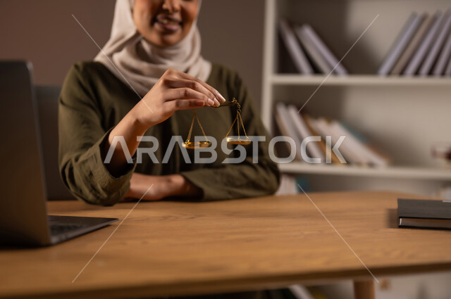 Work in the field of judiciary and law, the concept of justice and case study, legal professions and jobs, study of law and law, close-up portrait of a smiling Saudi Gulf Arab woman wearing a colorful abaya holding the scales of justice in her hand, use of modern devices and technologies in the legal field, beige background