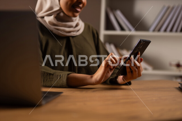 إجراء مكالمة فيديو عبر الجوال, تصفح الانترنت واستخدام تقنيات حديثة, الدردشة والمحادثة مع الأهل والاصدقاء, متابعة أخبار وسائل التواصل الاجتماعي, بورتريه مقرب  لإمرأة عربية خليجية سعودية مبتسمة ترتدي العباءة تستخدم الهاتف المحمول, خلفية بيج