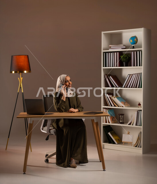 التواصل مع الأصدقاء عبر السوشال ميديا, الاستمتاع بتبادل الأحاديث والأخبار, إجراء مكالمة صوتية عبر الجوال, الدردشة والمحادثة عبر الأنترنت, بورتريه لشابة عربية خليجية سعودية محجبة مبتسمة ترتدي العباءة تستخدم الحاسب المحمول في المكتبة, خلفية بيج