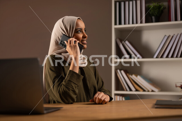 إجراء مكالمة صوتية عبر الجوال, الدردشة والمحادثة عبر الأنترنت, بورتريه مقرب لشابة عربية خليجية سعودية محجبة مبتسمة ترتدي العباءة تستخدم الحاسب المحمول في المكتبة, التواصل مع الأصدقاء عبر السوشال ميديا, الاستمتاع بتبادل الأحاديث والأخبار, خلفية بيج