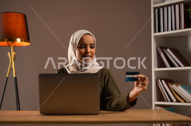 إمكانية تسديد الفواتير بالبطاقات الإلكترونية, الشراء والدفع عن طريق بطاقة الإئتمان البنكية، بورتريه مقرب لإمرأة عربية خليجية سعودية ترتدي العباءة تمسك بيدها بطاقة الصراف وتعمل على الحاسب المحمول, استخدام الأجهزة والتقنيات الحديثة في المكتبة, خلفية بيج