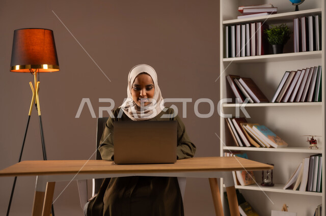 Using advanced devices and technologies, a portrait of a Saudi Gulf Arab woman wearing an abaya working on a computer, managing and completing business online, online shopping through applications and modern technology, conducting a remote job interview, browsing social networking sites, beige background