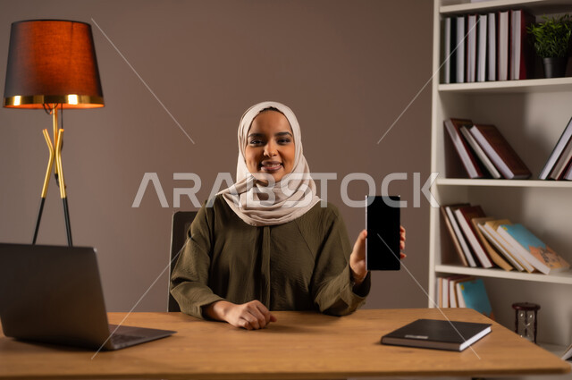 Displaying a blank black screen on a mobile phone with happiness gestures, using modern and advanced devices and technologies in the Kingdom’s offices, a portrait of a Saudi Gulf Arab woman wearing an abaya, using a laptop, browsing social media, beige background