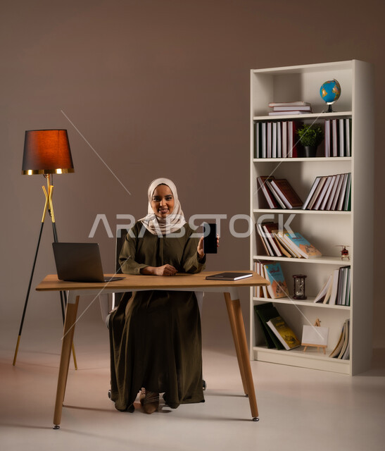 تصفح وسائل التواصل الاجتماعي, عرض شاشة سوداء فارغة عبر الهاتف المحمول بإيماءات السعادة، استخدام الأجهزة والتقنيات الحديثة والمتطورة في مكاتب المملكة, بورتريه لإمرأة عربية خليجية سعودية ترتدي العباءة تستخدم الحاسب المحمول, خلفية بيج