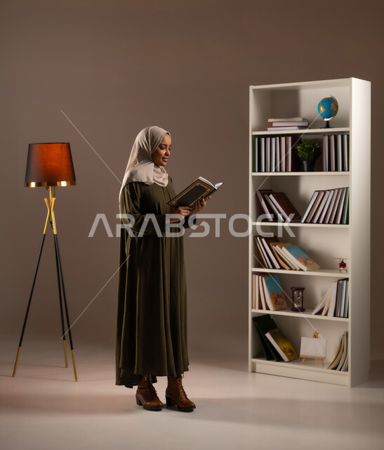 الاستمتاع بقراءة الكتب والقصص والروايات, ثقافة التعلم الذاتي, إنجاز الواجبات اليومية, قضاء الوقت بالقراءة والتعلم والتثقف، بورتريه مقرب من الجانب لامرأة عربية سعودية خليجية محجبة ترتدي العباءة تحمل بيديها كتاب، صورة الجسم بالطول الكامل, خلفية بيج