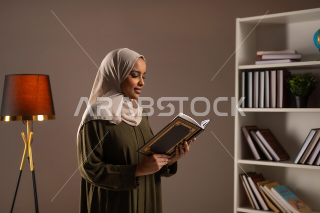 ثقافة التعلم الذاتي, إنجاز الواجبات اليومية, قضاء الوقت بالقراءة والتعلم والتثقف، بورتريه مقرب من الجانب لامرأة عربية سعودية خليجية محجبة ترتدي العباءة تحمل بيديها كتاب، الاستمتاع بقراءة الكتب والقصص والروايات, خلفية بيج
