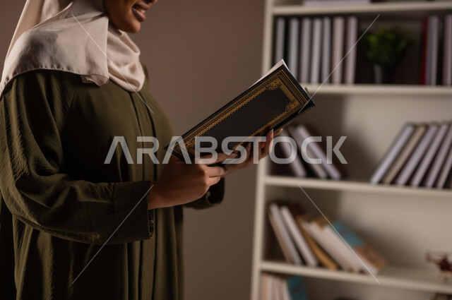 Completing daily duties, spending time reading, learning and acculturating, a side portrait of a veiled Saudi Arabian Gulf woman wearing an abaya, holding a book in her hands, enjoying reading books, stories and novels, self-learning culture, beige background