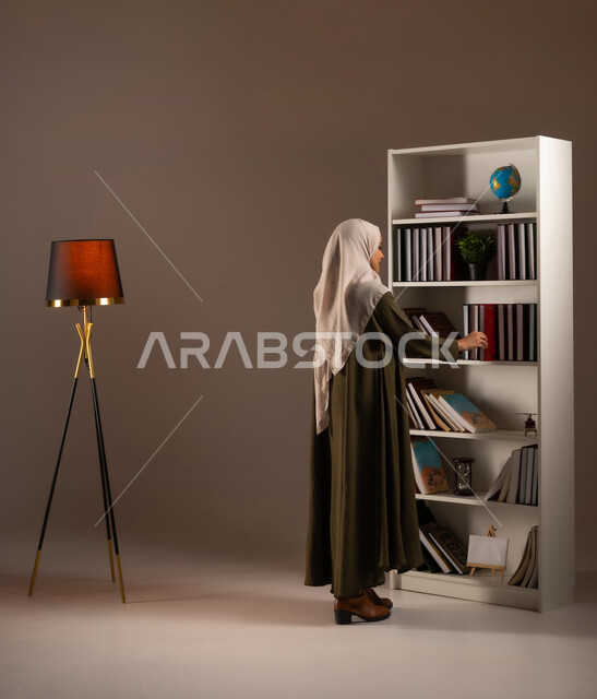 اتباع عادات وأسلوب حياة صحي, مفهوم القراءة والتثقيف الذاتي والتعلم, بورتريه من الجانب لشابة عربية خليجية سعودية ترتدي العباءة تقوم باقتناء كتاب من المكتبة, كتب الروايات والقصص, ممارسة الهوايات المفيدة, صورة الجسم بالكامل, خلفية بيج