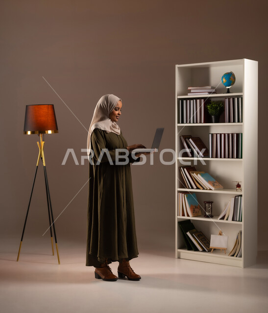 استخدام الأجهزة المتطورة في مكاتب المملكة, إدراة الأعمال عن بعد, الشراء والتسوق الالكتروني عبر التطبيقات والتقنيات الحديثة, بورتريه من الجانب لشابة عربية خليجية سعودية محجبة ترتدي العباءة تعمل على الحاسب المحمول, صورة الجسم بالطول الكامل, خلفية بيج