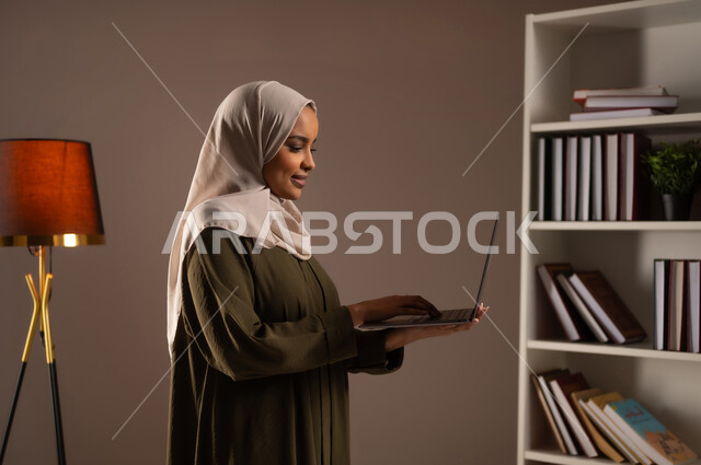 إنجاز وإدارة مهام العمل عبر الابتوب, استخدام الأجهزة المتطورة في مكاتب المملكة, الشراء والتسوق الالكتروني عبر التطبيقات والتقنيات الحديثة, بورتريه مقرب من الجانب لشابة عربية خليجية سعودية محجبة ترتدي العباءة تعمل على الحاسب المحمول, خلفية بيج