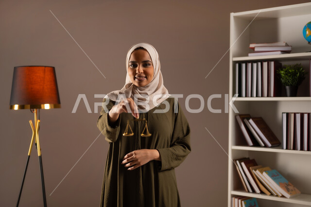 The concept of justice and case study, work in the field of judiciary and law, legal professions and jobs, study of law and law, close-up portrait of a smiling, veiled Saudi Arabian Gulf woman wearing a colorful abaya, holding the scales of justice in her hand, beige background