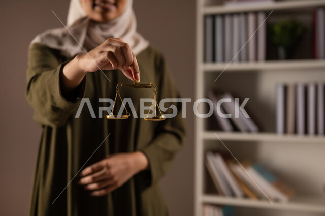 Work in the field of judiciary and law, the concept of justice and case study, legal professions and jobs, study of law and law, portrait of a smiling Saudi Gulf Arab woman wearing a veiled abaya, holding the scales of justice in her hand, beige background