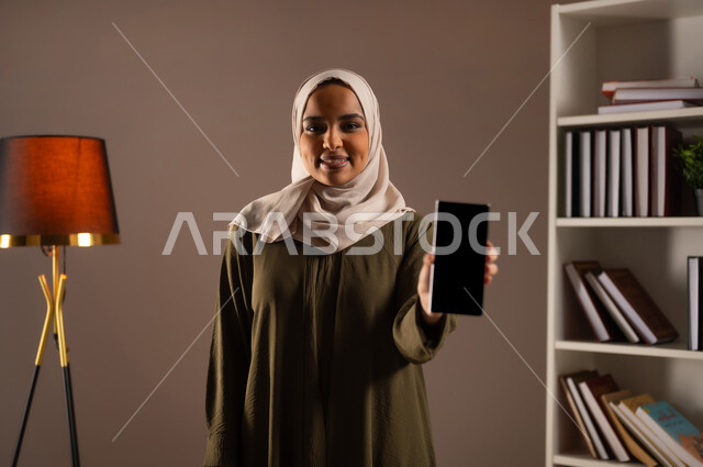 عرض شاشة سوداء فارغة عبر الجوال بإيماءات السعادة، استخدام الأجهزة والتقنيات الحديثة والمتطورة في مكاتب المملكة, بورتريه مقرب لإمرأة عربية خليجية سعودية ترتدي العباءة تستخدم تطبيقات وخدمات الهاتف المحمول, تصفح وسائل التواصل الاجتماعي, خلفية بيج