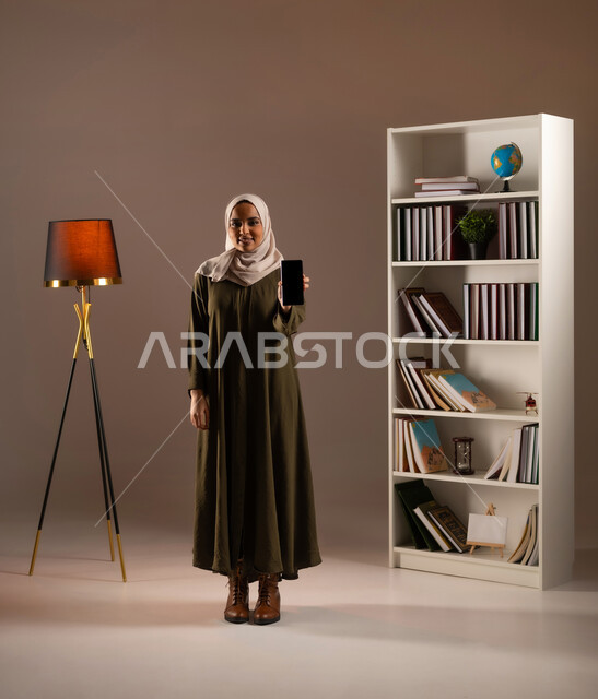 Browsing social media, displaying a blank black screen on the mobile phone, using modern and advanced devices and technologies in the Kingdom’s offices, a close-up portrait of a Saudi Gulf Arab woman wearing an abaya, using mobile phone applications and services, full body image, beige background