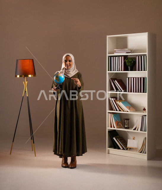 The concept of research and exploration around the world, pointing to a specific place on the map and looking at the camera with a smile, portrait of a Saudi Gulf Arab woman wearing a colorful abaya, holding a model of the Earth in the library in her hand, full-length body portrait, beige background