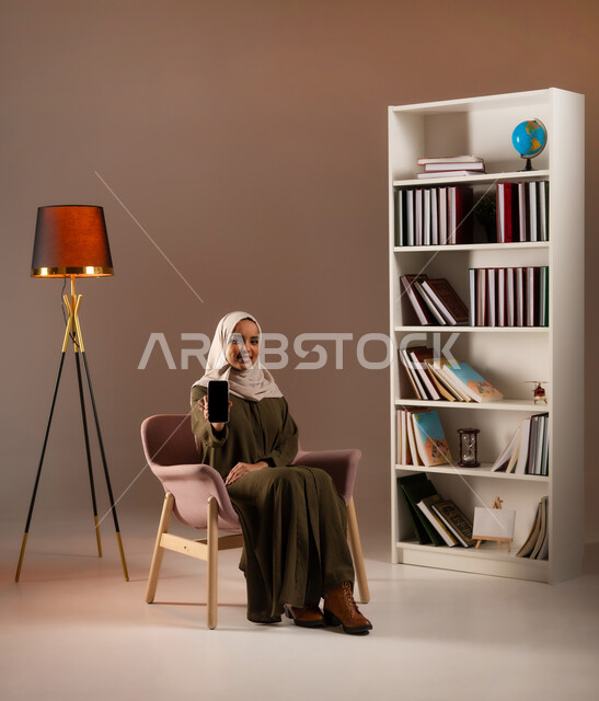 Using modern and advanced devices and technologies in the Kingdom’s offices, displaying a blank black screen on the mobile phone with happiness gestures, a portrait of a Saudi Gulf Arab woman wearing the abaya, using mobile phone applications and services, browsing social media, beige background