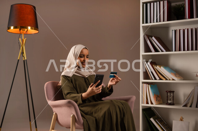 Browsing banking and banking applications via mobile phone, purchasing and paying remotely via the Internet, using a credit card in electronic shopping, a portrait of a Saudi Gulf Arab woman wearing an abaya, sitting on a chair and holding a bank card in the library, beige background