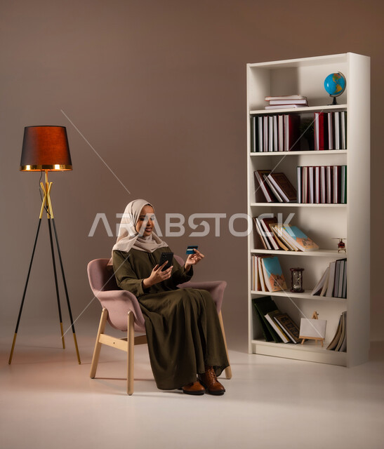 الشراء والدفع عن بعد عبر الاونلاين, استخدام بطاقة الإئتمان في التسوق الالكتروني, بورتريه لشابة عربية خليجية سعودية ترتدي العباءة تجلس على كرسي وتمسك البطاقة البنكية في المكتبة, تصفح تطبيقات المصارف والبنوك عبر الهاتف المحمول, خلفية بيج