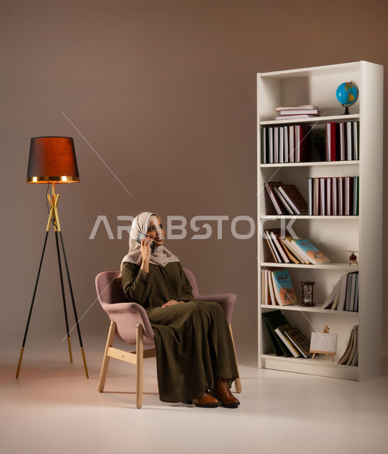 Enjoying the exchange of conversations and news, making a voice call via mobile phone, chatting and conversing over the Internet, a portrait of a veiled Saudi Gulf Arab woman, smiling, wearing an abaya, using the mobile phone in the library, communicating with friends via social networking sites, beige background