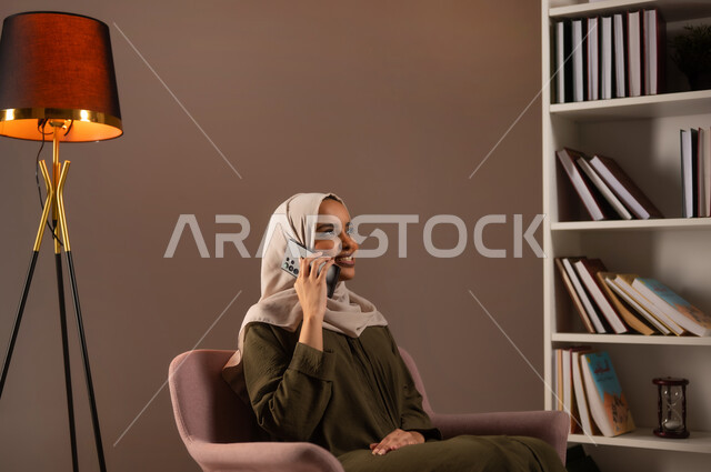 إجراء مكالمة صوتية عبر الجوال, الدردشة والمحادثة عبر الأنترنت, بورتريه لشابة عربية خليجية سعودية محجبة مبتسمة ترتدي العباءة تستخدم الهاتف المحمول في المكتبة, التواصل مع الأصدقاء عبر السوشال ميديا, الاستمتاع بتبادل الأحاديث والأخبار, خلفية بيج