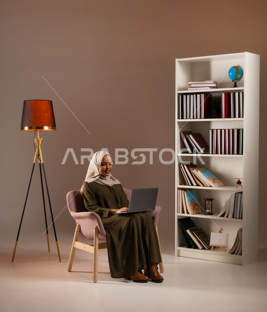 Using advanced devices in the Kingdom’s offices, buying books and online shopping through modern applications and technologies, a portrait of a smiling Saudi Gulf Arab woman wearing an abaya, working on a laptop, managing business remotely, beige background