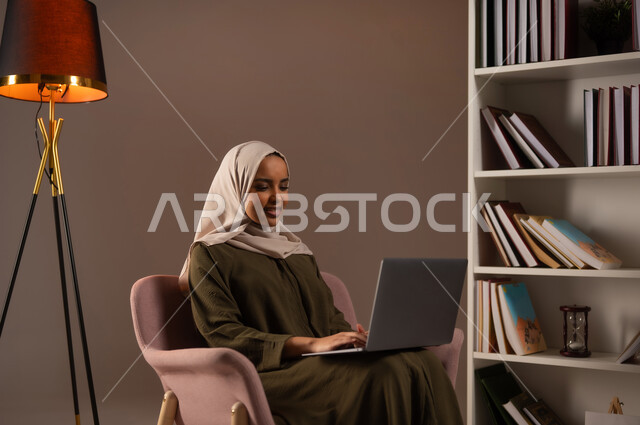 شراء الكتب والتسوق الالكتروني عبر التطبيقات والتقنيات الحديثة, بورتريه لشابة عربية خليجية سعودية مبتسمة محجبة ترتدي العباءة تعمل على الحاسب المحمول, استخدام الأجهزة المتطورة في مكاتب المملكة, إدراة الأعمال عن بعد, خلفية بيج