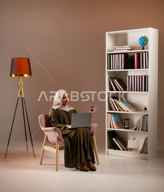 استخدام الأجهزة والتقنيات الحديثة في المكتبة, الشراء والدفع عن طريق بطاقة الإئتمان البنكية، بورتريه لإمرأة عربية خليجية سعودية ترتدي العباءة تمسك بيدها بطاقة الصراف وتعمل على الحاسب المحمول, إمكانية تسديد الفواتير بالبطاقات الإلكترونية, خلفية بيج