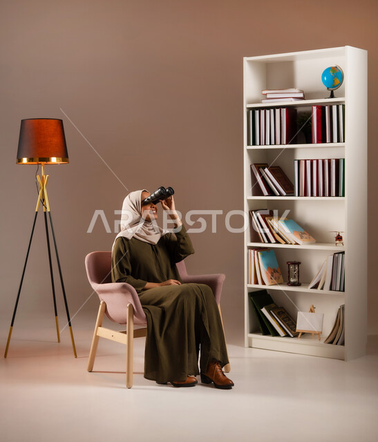 حب الاستكشاف والمغامرة, رؤية الاشياء البعيدة عبر المنظار بإيماءات السرور, التخطيط المستقبلي للأهداف و الطموحات، بورتريه لإمرأة عربية خليجية سعودية ترتدي العباءة تقوم بالنظر عبر منظار ثنائي العينين (التلسكوب) في المكتبة, خلفية بيج