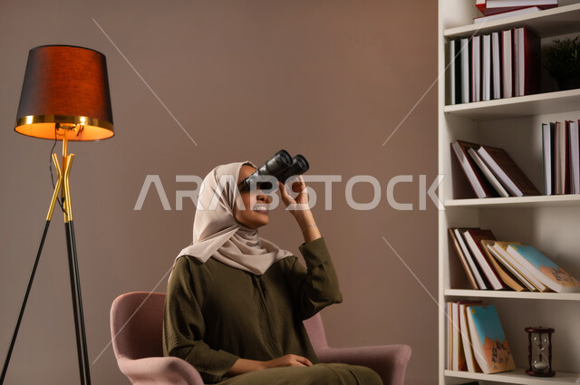 رؤية الاشياء البعيدة عبر المنظار بإيماءات السرور, التخطيط المستقبلي للأهداف و الطموحات، حب الاستكشاف والمغامرة, بورتريه لإمرأة عربية خليجية سعودية ترتدي العباءة تقوم بالنظر عبر منظار ثنائي العينين (التلسكوب) في المكتبة, خلفية بيج