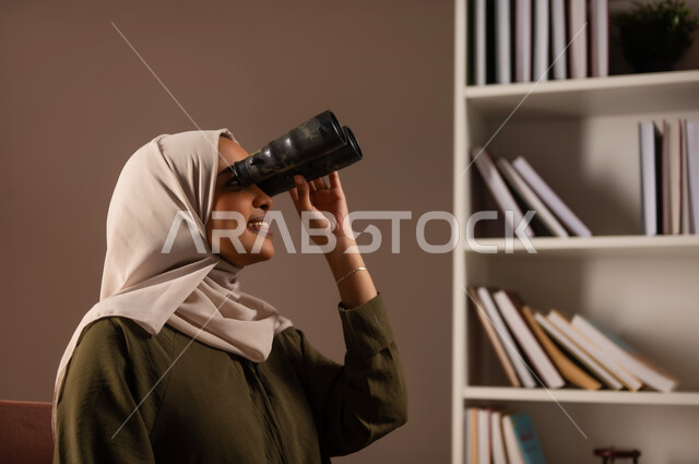 التخطيط المستقبلي للأهداف و الطموحات، رؤية الاشياء البعيدة عبر المنظار بإيماءات السرور، حب الاستكشاف والمغامرة, بورتريه مقرب من الجانب لإمرأة عربية خليجية سعودية ترتدي العباءة تقوم بالنظر عبر منظار ثنائي العينين (التلسكوب) في المكتبة, خلفية بيج