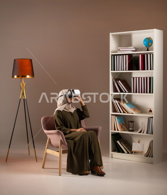 الجلوس بتركيز ومشاهدة أفلام الواقع الافتراضي, التفاعل والاندماج ومحاكاة العالم الخيالي, بورتريه لشابة عربية خليجية سعودية ترتدي العباءة الملونة ونظارة في آر ثلاثية الأبعاد تجلس على الكرسي في المكتبة, الترفيه والتسلية والاستمتاع في وقت الفراغ, خلفية بيج